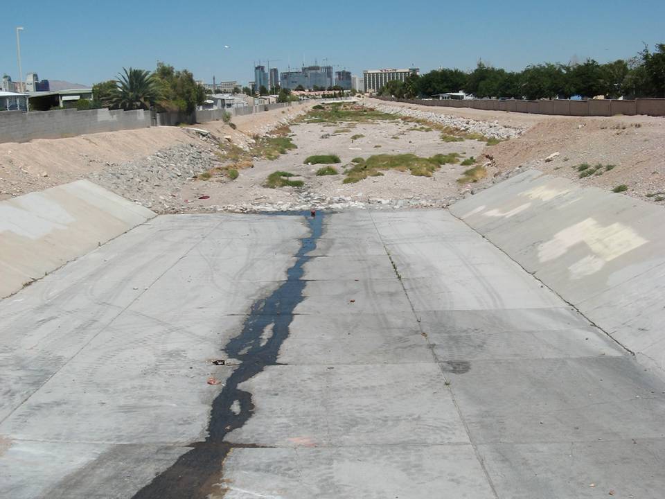 A wide dry concrete flood channel with a dark wet strip down the middle, desert gravel banks and the Strip skyline behind