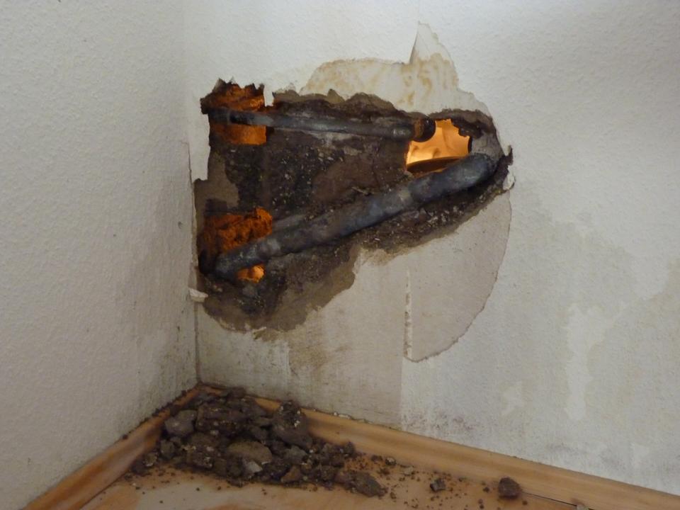 Plaster wall broken open around corroded pipes, with crumbled plaster on the floor